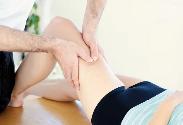 Physiotherapeut untersucht das angewinkelte Knie einer auf einer Liege sitzenden Person.