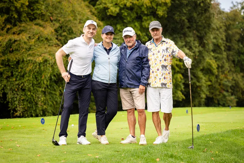 Vier Personen stehen mit Golfschlägern auf einem Golfplatz vor dichtem Baumbestand.