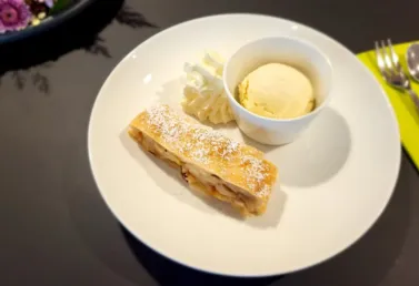 Apfelstrudel mit Puderzucker, serviert mit einer Portion Vanilleeis und einem Klecks Schlagsahne auf einem weißen Teller, daneben ein grünes Serviettenstück und Besteck.