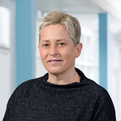 Porträt einer Person mit kurzem blondem Haar und schwarzem Oberteil vor hellem Hintergrund mit blauen Fensterrahmen.