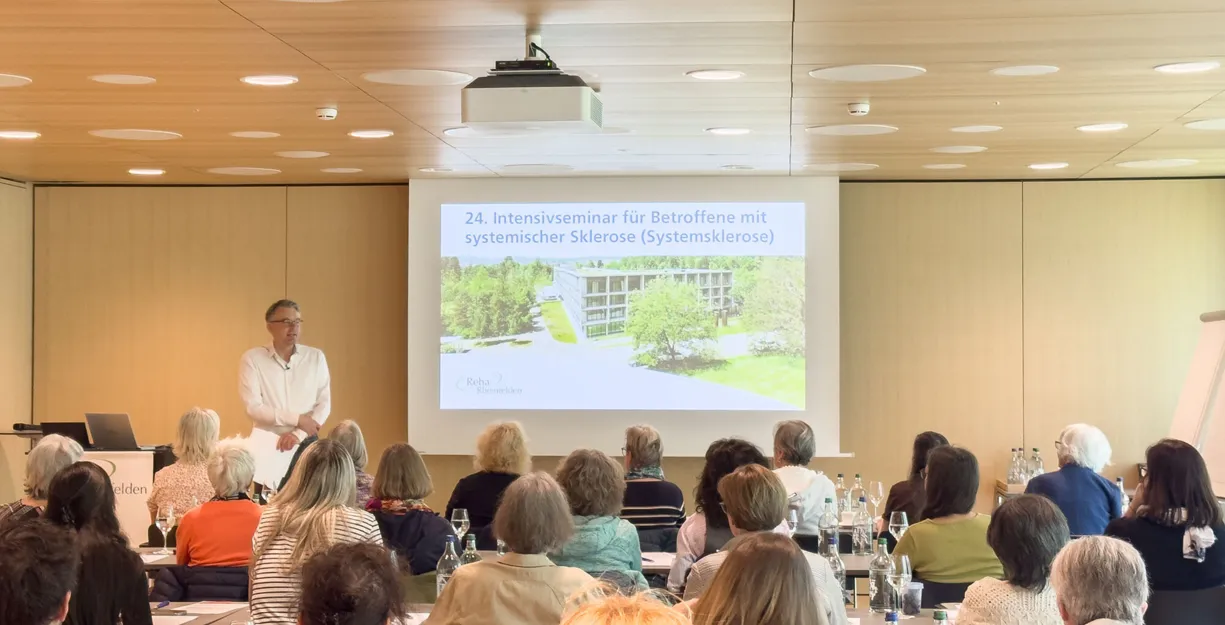 Seminarraum mit Publikum an Tischen, Sprecher steht vorne und hält eine Präsentation auf einer Leinwand mit Text „24. Intensivseminar für Betroffene…“ und Gebäudeansicht im Hintergrund.