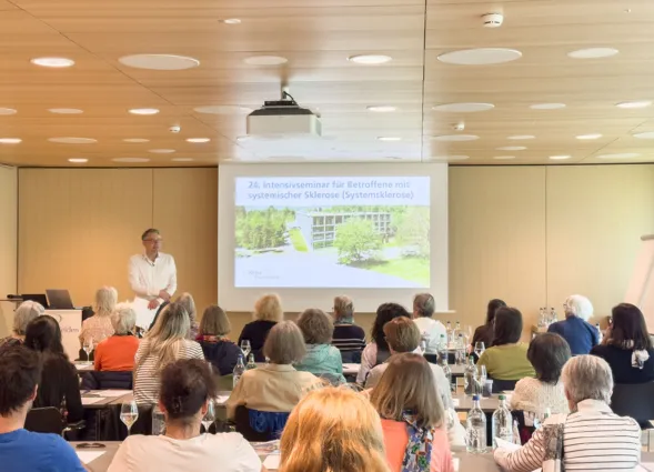 Seminarraum mit Publikum an Tischen, Sprecher steht vorne und hält eine Präsentation auf einer Leinwand mit Text „24. Intensivseminar für Betroffene…“ und Gebäudeansicht im Hintergrund.