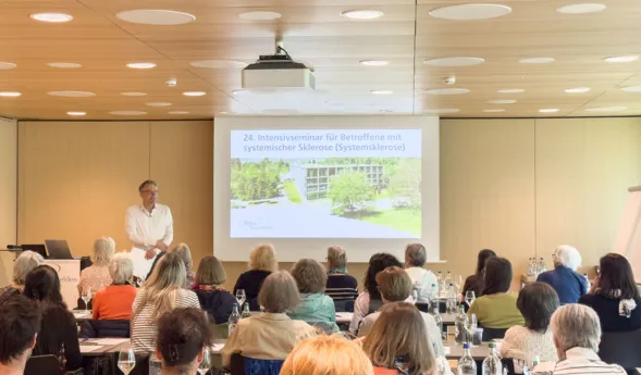Seminarraum mit Publikum an Tischen, Sprecher steht vorne und hält eine Präsentation auf einer Leinwand mit Text „24. Intensivseminar für Betroffene…“ und Gebäudeansicht im Hintergrund.
