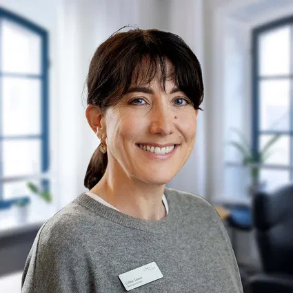 Lächelnde Frau mit dunklem Haar und grauem Pullover, Namensschild an der Brust, in einem hellen Büro mit großen Fenstern.