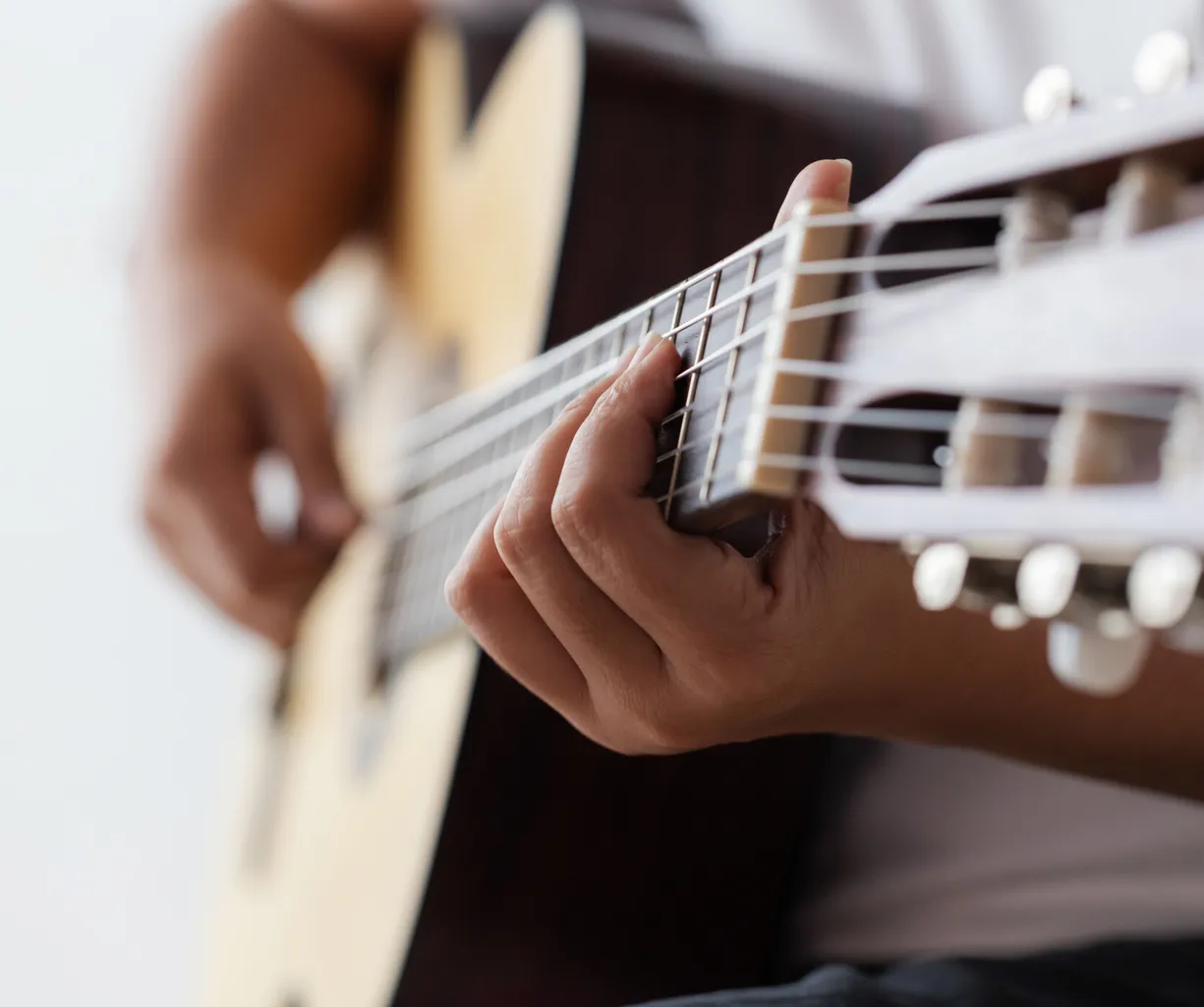 Nahaufnahme einer Hand, die eine Gitarre spielt. Die Finger drücken die Saiten, während die andere Hand den Hals der Gitarre hält. Der Hintergrund ist unscharf und hell, was den Fokus auf die Hände und das Instrument lenkt.