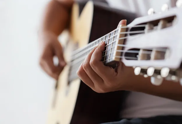 Nahaufnahme einer Hand, die eine Gitarre spielt. Die Finger drücken die Saiten, während die andere Hand den Hals der Gitarre hält. Der Hintergrund ist unscharf und hell, was den Fokus auf die Hände und das Instrument lenkt.