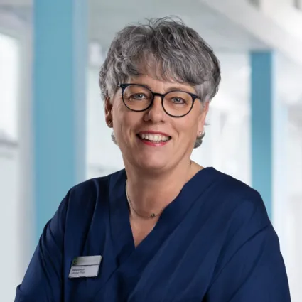 Lächelnde Frau mit kurzen grauen Haaren, Brille und blauer Arbeitskleidung, steht vor einem hellen, modernen Hintergrund. Ein Namensschild ist sichtbar.