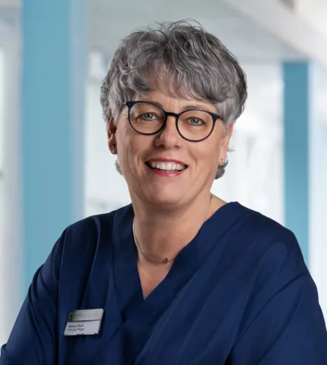 Lächelnde Frau mit kurzen grauen Haaren, Brille und blauer Arbeitskleidung, steht vor einem hellen, modernen Hintergrund. Ein Namensschild ist sichtbar.