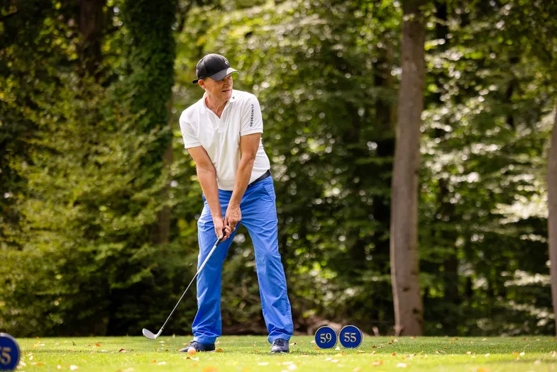 Golfspieler mit schwarzer Kappe, weißem Shirt und blauer Hose schlägt im Wald auf einem Rasenplatz; daneben stehen blaue Markierungen mit den Zahlen 59 und 55.
