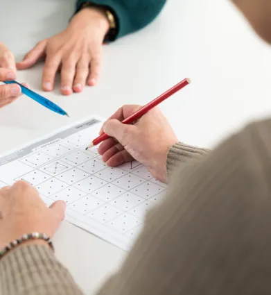 Zwei Hände arbeiten an einem Arbeitsblatt mit Punkten und Linien. Eine Hand hält einen roten Bleistift und zeichnet auf dem Blatt, während die andere Hand einen blauen Stift hält und auf das Blatt zeigt.