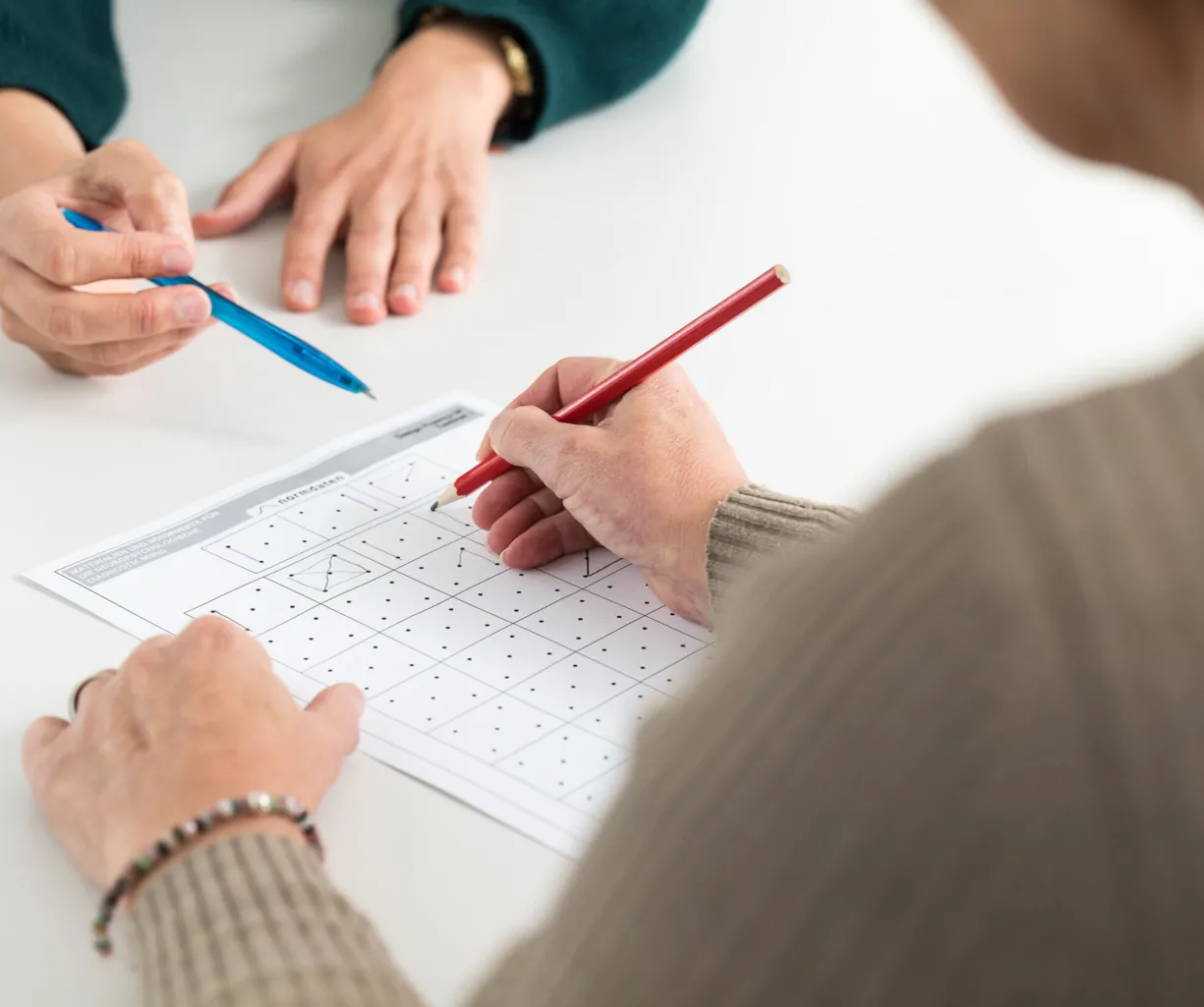 Zwei Hände arbeiten an einem Arbeitsblatt mit Punkten und Linien. Eine Hand hält einen roten Bleistift und zeichnet auf dem Blatt, während die andere Hand einen blauen Stift hält und auf das Blatt zeigt.
