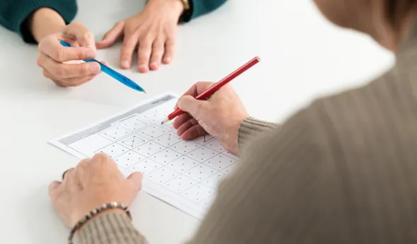 Zwei Hände arbeiten an einem Arbeitsblatt mit Punkten und Linien. Eine Hand hält einen roten Bleistift und zeichnet auf dem Blatt, während die andere Hand einen blauen Stift hält und auf das Blatt zeigt.
