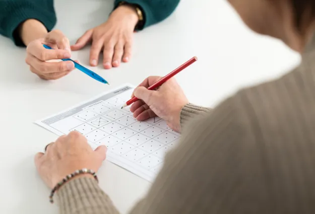 Zwei Hände arbeiten an einem Arbeitsblatt mit Punkten und Linien. Eine Hand hält einen roten Bleistift und zeichnet auf dem Blatt, während die andere Hand einen blauen Stift hält und auf das Blatt zeigt.