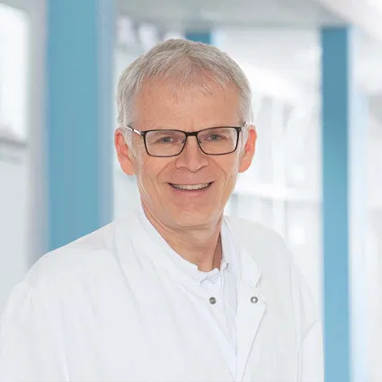 Portrait eines älteren Mannes mit grauem Haar, Brille und weißem Kittel vor blau-weißem Hintergrund, freundlich lächelnd.