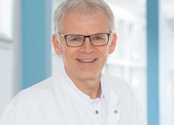 Portrait eines älteren Mannes mit grauem Haar, Brille und weißem Kittel vor blau-weißem Hintergrund, freundlich lächelnd.