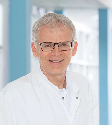 Portrait eines älteren Mannes mit grauem Haar, Brille und weißem Kittel vor blau-weißem Hintergrund, freundlich lächelnd.