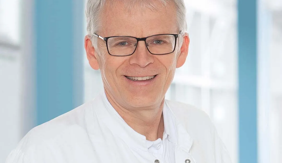 Portrait eines älteren Mannes mit grauem Haar, Brille und weißem Kittel vor blau-weißem Hintergrund, freundlich lächelnd.