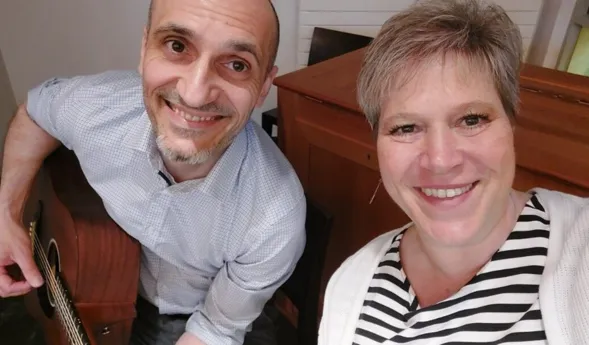 Lächelnde Frau mit kurzen, grauen Haaren und gestreiftem Oberteil sitzt neben einem Mann mit Glatze und Bart, der eine Gitarre hält. Im Hintergrund ist ein Holztisch sichtbar.