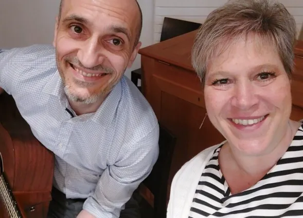 Lächelnde Frau mit kurzen, grauen Haaren und gestreiftem Oberteil sitzt neben einem Mann mit Glatze und Bart, der eine Gitarre hält. Im Hintergrund ist ein Holztisch sichtbar.