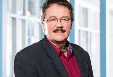 Mann mit Brille, Schnurrbart und grauen Haaren, trägt einen dunklen Blazer über einem roten Hemd, steht vor einer hellen, modernen Hintergrundarchitektur mit großen Fenstern.