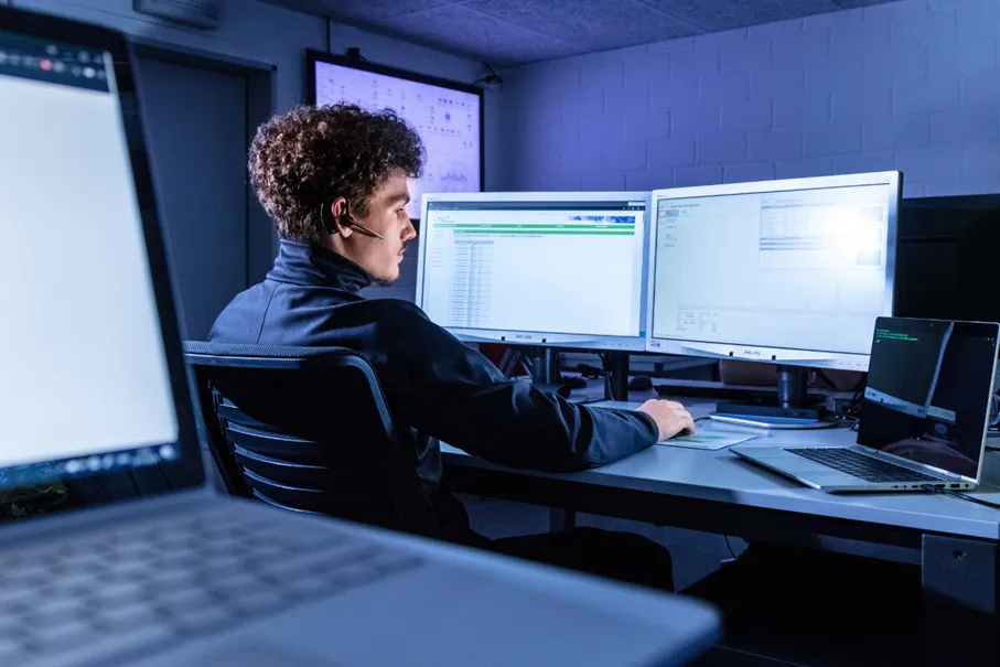 Mann mit Headset sitzt an einem Schreibtisch, umgeben von mehreren Monitoren, die Daten anzeigen. Ein Laptop ist ebenfalls auf dem Tisch platziert. Die Umgebung ist schwach beleuchtet, mit einem blauen Lichtakzent.