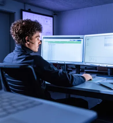 Mann mit Headset sitzt an einem Schreibtisch, umgeben von mehreren Monitoren, die Daten anzeigen. Ein Laptop ist ebenfalls auf dem Tisch platziert. Die Umgebung ist schwach beleuchtet, mit einem blauen Lichtakzent.