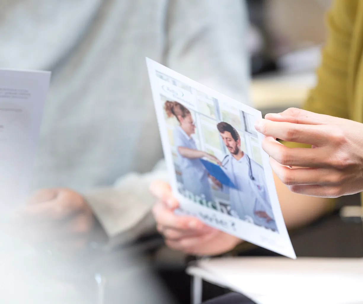 Eine Person hält ein Informationsblatt mit einem Bild von zwei medizinischen Fachkräften in einem hellen Raum. Im Hintergrund sind weitere Dokumente und Personen teilweise sichtbar.