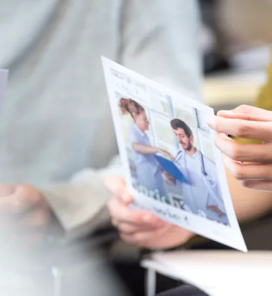 Eine Person hält ein Informationsblatt mit einem Bild von zwei medizinischen Fachkräften in einem hellen Raum. Im Hintergrund sind weitere Dokumente und Personen teilweise sichtbar.