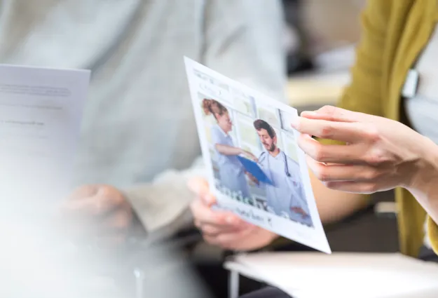 Eine Person hält ein Informationsblatt mit einem Bild von zwei medizinischen Fachkräften in einem hellen Raum. Im Hintergrund sind weitere Dokumente und Personen teilweise sichtbar.