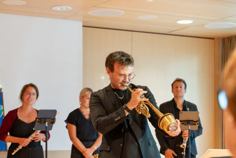 Musiker spielen in einem Innenraum vor Publikum; ein Trompeter steht im Mittelpunkt, begleitet von weiteren Bläsern und einem Kontrabass.