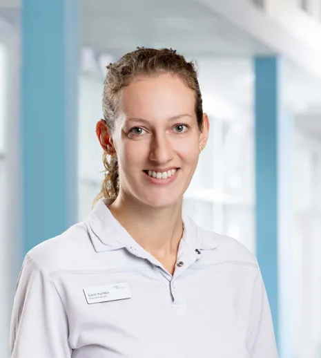 Lächelnde Frau mit lockigem Haar, trägt ein weißes Polohemd und ein Namensschild, vor einem hellen, modernen Hintergrund mit blauen Akzenten.