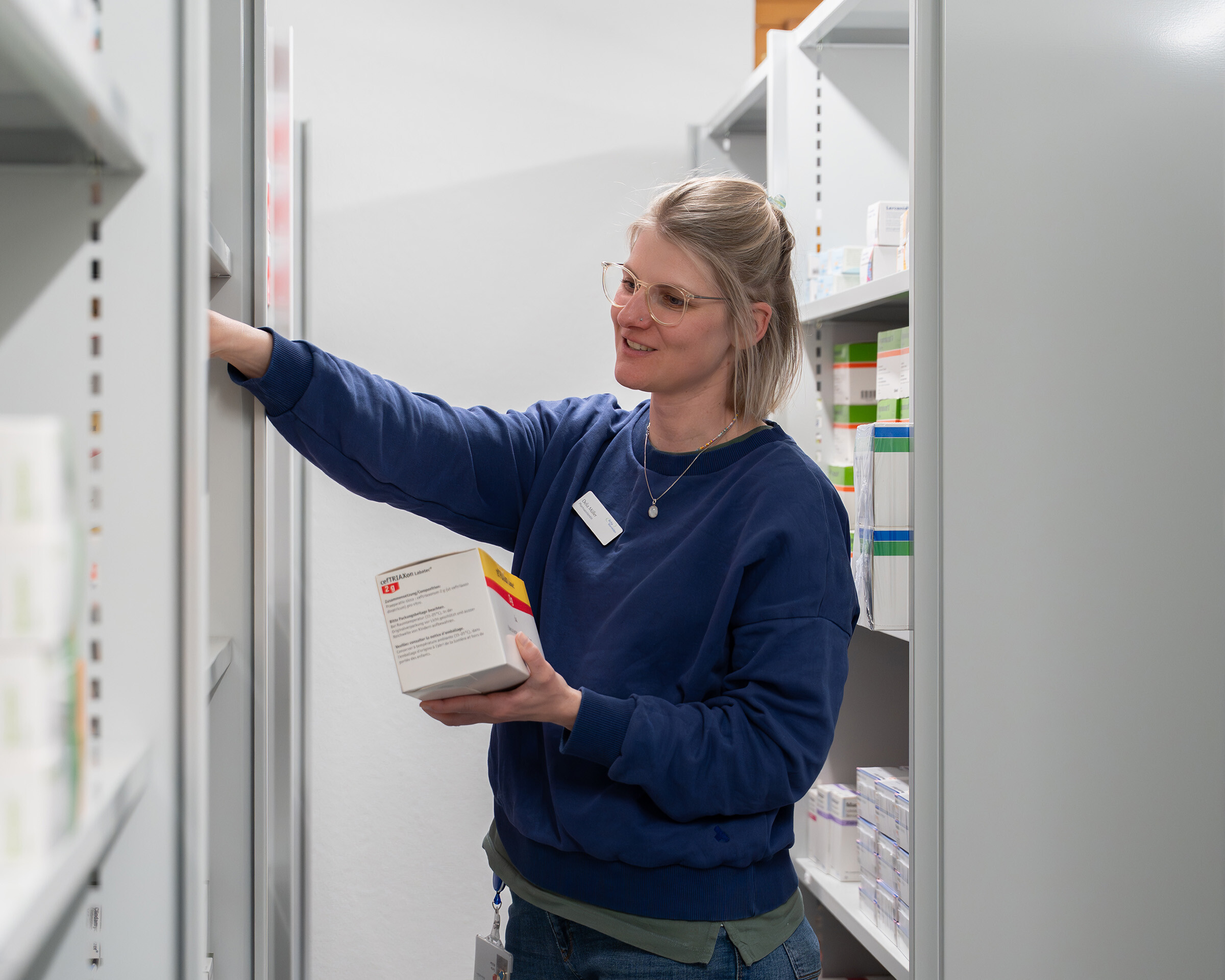 Mitarbeiterin in blauem Pullover entnimmt eine Medikamentenschachtel aus einem Apothekenregal.