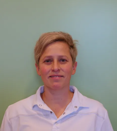 Porträt einer Person mit kurzen, blonden Haaren, die ein weißes Oberteil trägt, vor einer grünen Wand. Die Person lächelt leicht und blickt direkt in die Kamera.