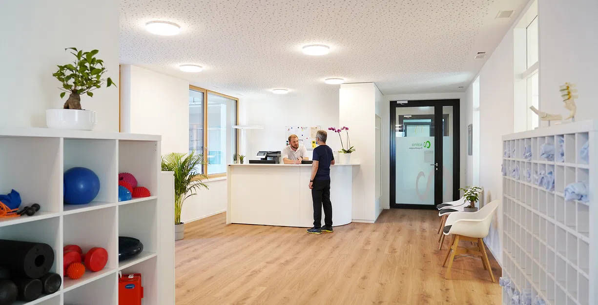 Heller Empfangsbereich einer Praxis mit Rezeption, zwei Personen, Trainingsgeräten im Regal, Sitzgelegenheiten und Schließfächern.