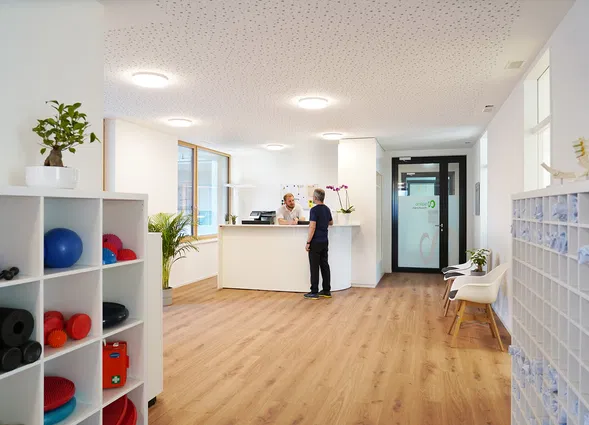 Heller Empfangsbereich einer Praxis mit Rezeption, zwei Personen, Trainingsgeräten im Regal, Sitzgelegenheiten und Schließfächern.