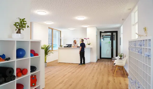 Heller Empfangsbereich einer Praxis mit Rezeption, zwei Personen, Trainingsgeräten im Regal, Sitzgelegenheiten und Schließfächern.