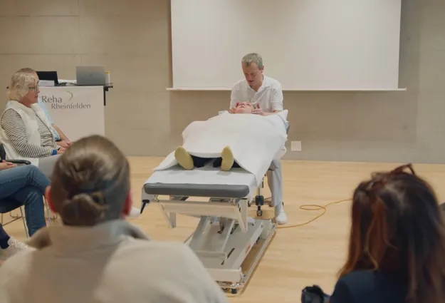 Ein Therapeut führt eine Behandlung an einer liegenden Person auf einer Behandlungsliege durch, während eine Gruppe von Zuschauern in einem Seminarraum aufmerksam zuschaut. Im Hintergrund ist ein Präsentationsbildschirm und ein Tisch mit einem Laptop s...