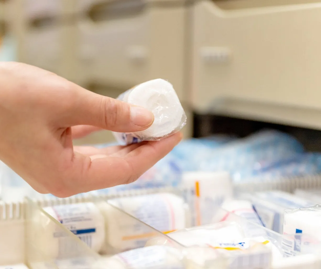 Hand hält eine verpackte medizinische Rolle über einem Fach mit weiteren medizinischen Produkten in einer Apotheke.