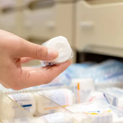 Hand hält eine verpackte medizinische Rolle über einem Fach mit weiteren medizinischen Produkten in einer Apotheke.