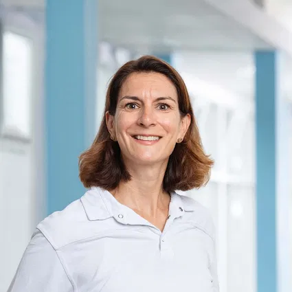 Lächelnde Frau mit schulterlangem braunem Haar und weißem Hemd vor hellem, modernem Innenraum.
