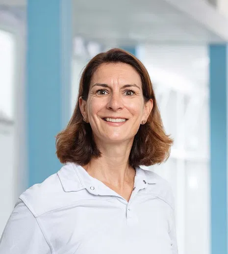 Lächelnde Frau mit schulterlangem braunem Haar und weißem Hemd vor hellem, modernem Innenraum.