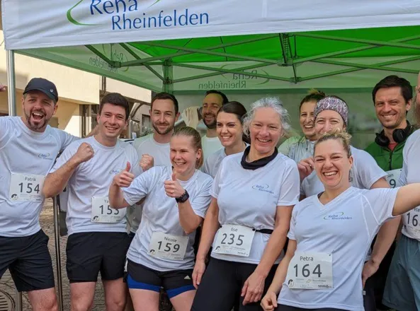 Gruppe von Läufern in weißen T-Shirts mit Startnummern, lächelnd und posierend unter einem grünen Zelt mit dem Logo „Reha Rheinfelden“. Im Hintergrund sind weitere Personen und eine Gebäudewand sichtbar.