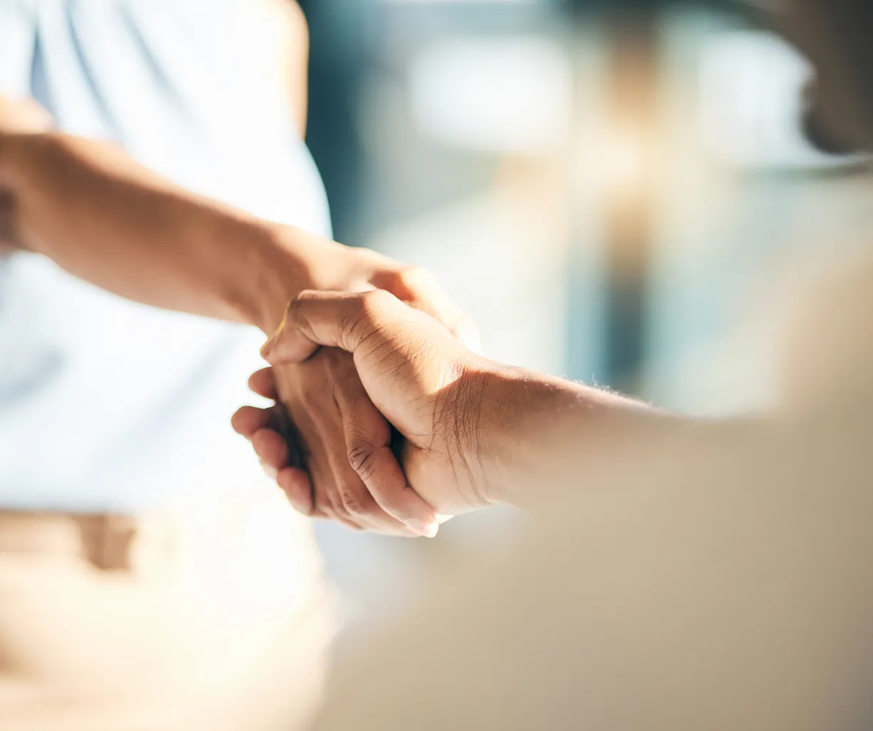 Zwei Hände, die sich in einem festen Händedruck treffen, im Hintergrund unscharf. Die Hände sind unterschiedlich gefärbt und zeigen eine freundliche Interaktion.