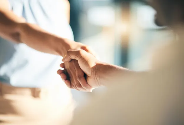 Zwei Hände, die sich in einem festen Händedruck treffen, im Hintergrund unscharf. Die Hände sind unterschiedlich gefärbt und zeigen eine freundliche Interaktion.