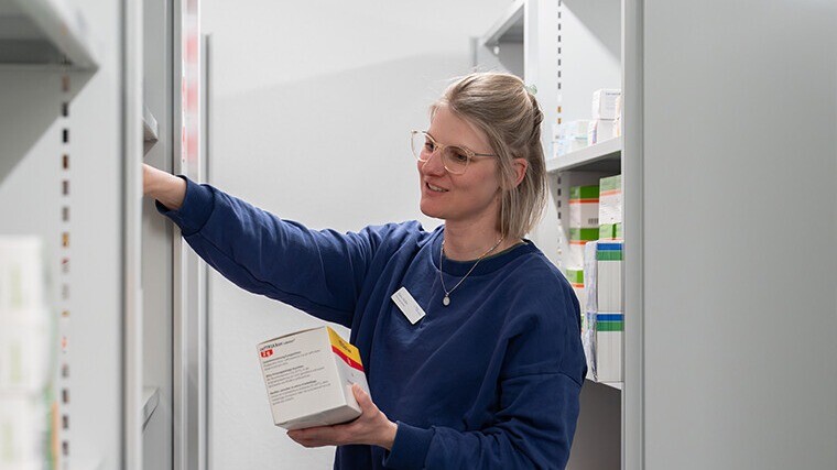 Mitarbeiterin in dunklem Outfit holt mit Buch/Ordner Dokumente aus einem offenen Lagerregal; im Hintergrund weitere Boxen und Ordner.