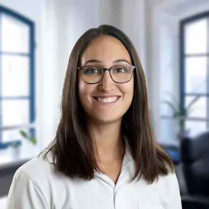 Lächelnde Frau mit langen braunen Haaren und Brille, in weißer Bluse vor hellen Bürofenstern.