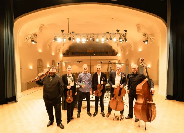 Sechs Musiker stehen auf einer Bühne in einem Konzertsaal. Sie halten verschiedene Instrumente, darunter Geigen, Bratsche und ein Cello. Der Saal hat eine elegante Architektur mit sanften Lichtquellen und einem Holzfußboden. Im Hintergrund sind weitere...
