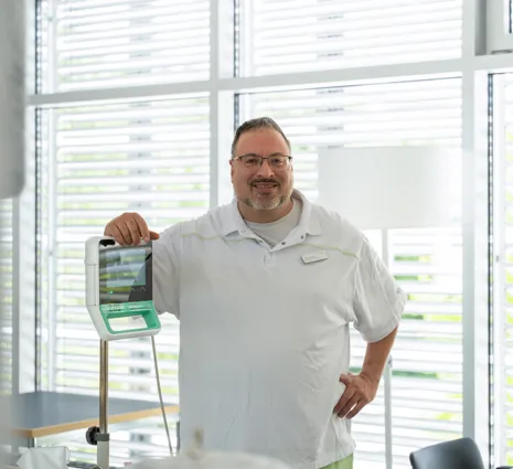 Mann im weißen Polohemd steht im Büro vor Fenstern mit Rollläden und hält ein Krankenhaus- bzw. medizinisches Gerät mit Griffen und Display.