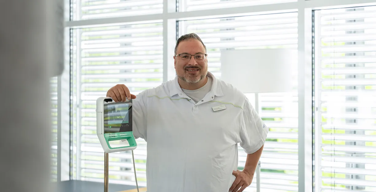 Mann im weißen Polohemd steht im Büro vor Fenstern mit Rollläden und hält ein Krankenhaus- bzw. medizinisches Gerät mit Griffen und Display.