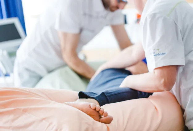 Zwei Personen in weißen Arbeitskleidung führen eine Behandlung an einem Patienten durch, der auf einer Liege mit einem rosa Laken liegt. Ein Fuß des Patienten ist sichtbar. Im Hintergrund sind medizinische Geräte und ein Vorhang zu sehen.